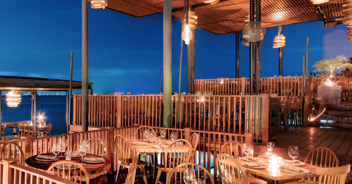 Multi-level outdoor restaurant with wood furniture, candlelit tables, and water views at dusk creates a warm, inviting mood.