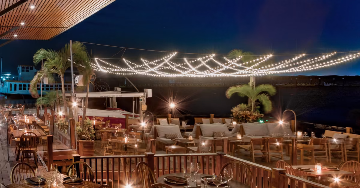 Nighttime waterfront restaurant with string lights, wooden seating, palm trees, cozy vibes, and a boat docked nearby.