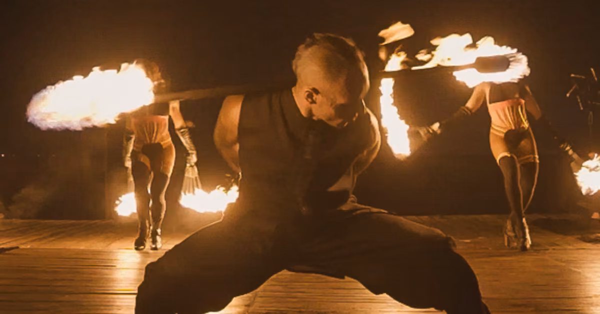 Three performers in dark outfits spin flaming batons on a dim stage; one in front bends low, staff spinning behind their back.
