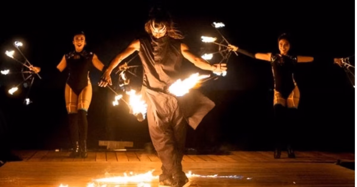 Three performers in dark outfits do a fire dance at Rosa Negra, with flaming props illuminating the stage’s dark backdrop.
