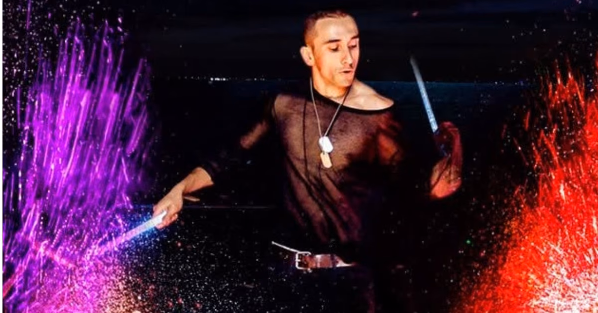 A person in a sheer black shirt holds glowing sticks, casting vibrant purple and red lights against a lively, dark background.
