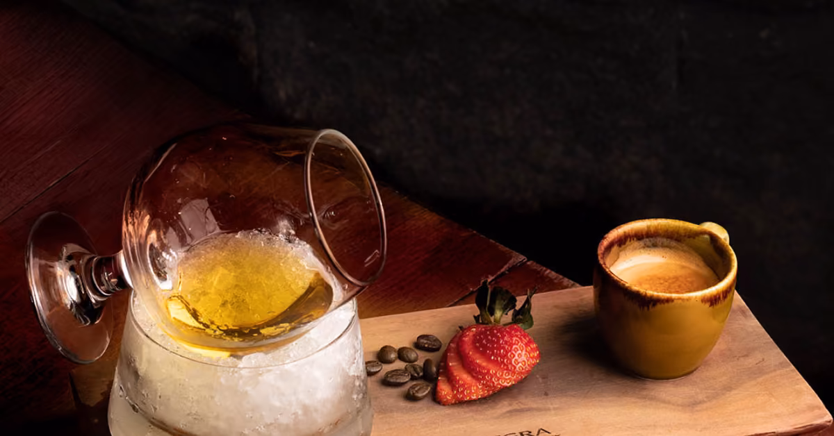 A sideways cocktail glass on ice, with espresso, coffee beans, and a sliced strawberry on a wooden board against a dark background.