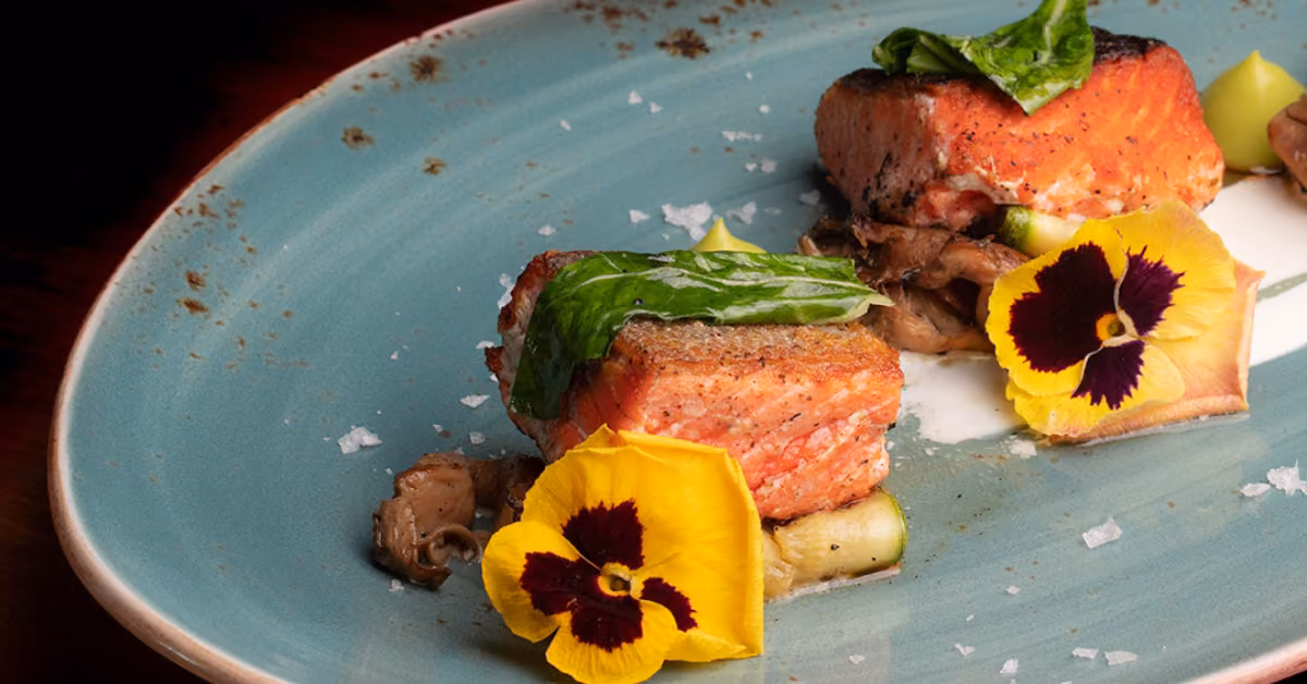 Seared salmon fillets topped with green leaves and yellow flowers, served with mushrooms on a blue plate.