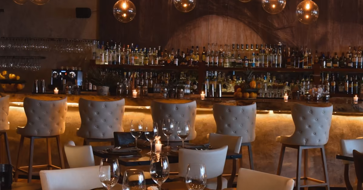 A dimly lit bar at Rosa Negra with high stools, globe lights, and tables set with glasses and candles creates a cozy vibe.