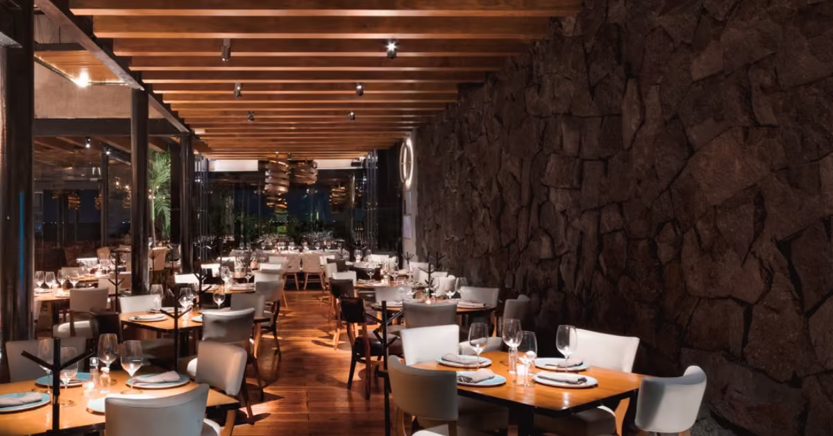 Modern restaurant with wooden floors, white chairs, stone wall, exposed beams, and warm lighting for an inviting atmosphere.