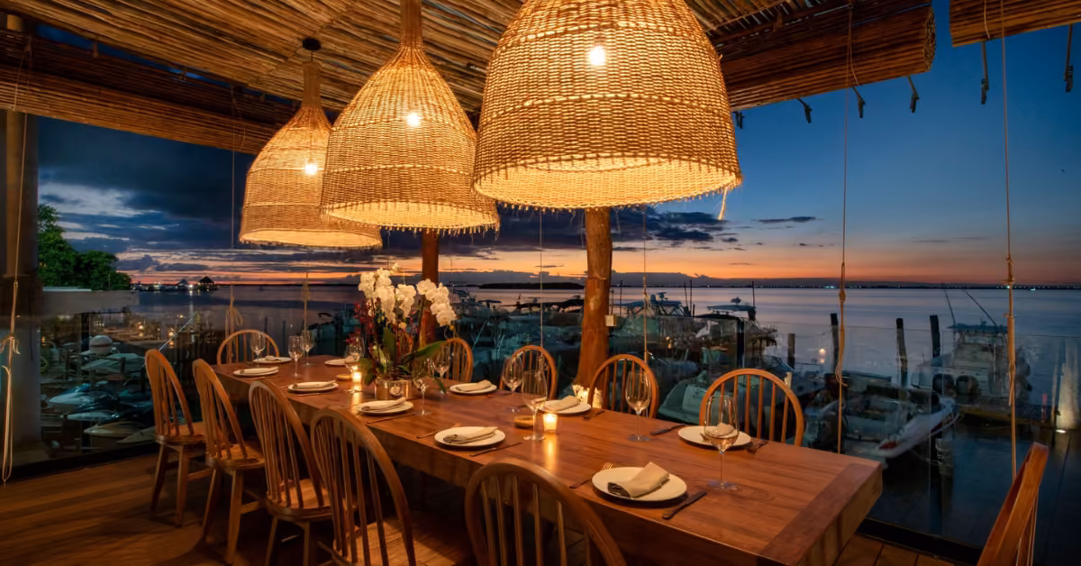 Sunset view from a waterfront restaurant with a set wooden table, wicker lights, and windows overlooking boats on calm water.