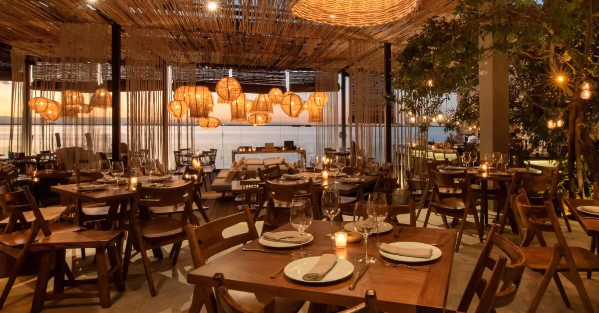 Warm, inviting restaurant with wood tables, wicker lamps, and large windows facing sunset-lit water; tables set for dining.