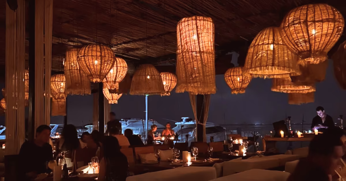 A cozy restaurant with woven basket lights casts a warm glow over candlelit tables as boats drift by outside at night.