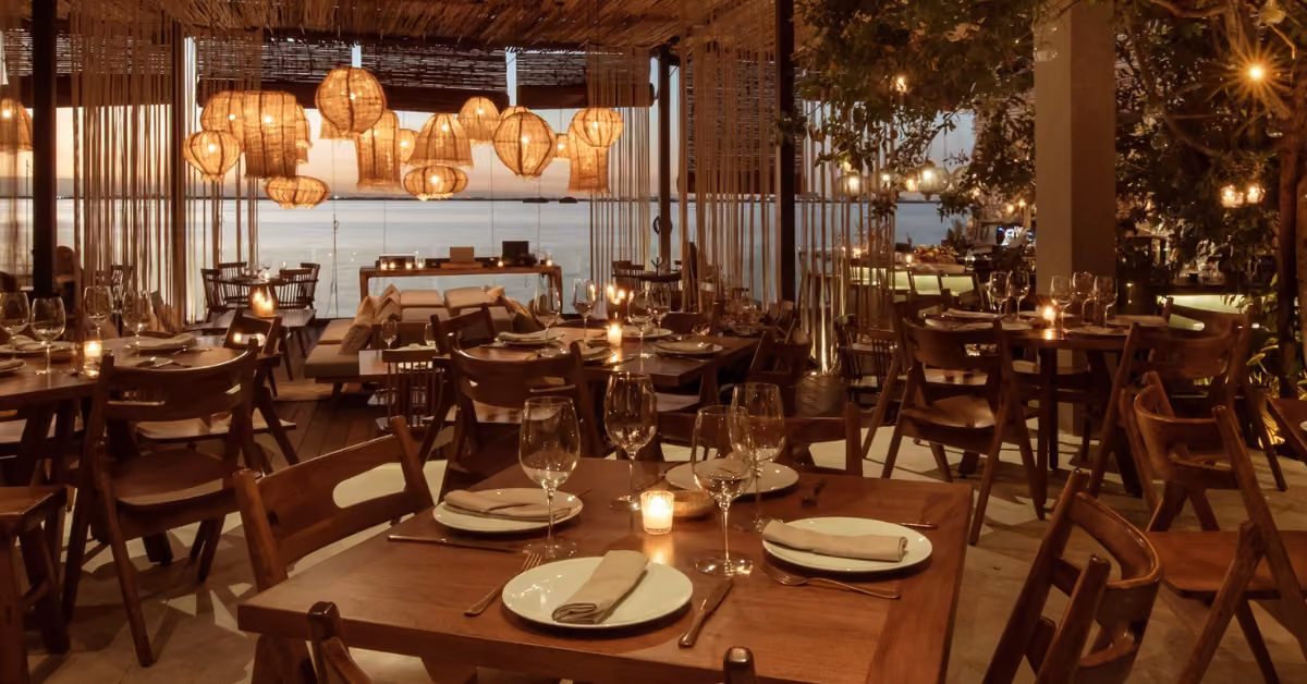 A warmly lit restaurant at sunset with wooden tables, candles, and woven lights overlooks the ocean in a cozy setting.