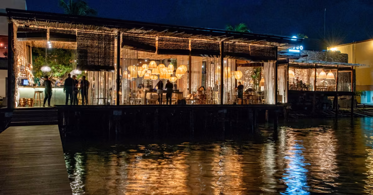 A modern glass-walled restaurant with warm lighting sits over water at night, its lights reflecting on the rippling surface.