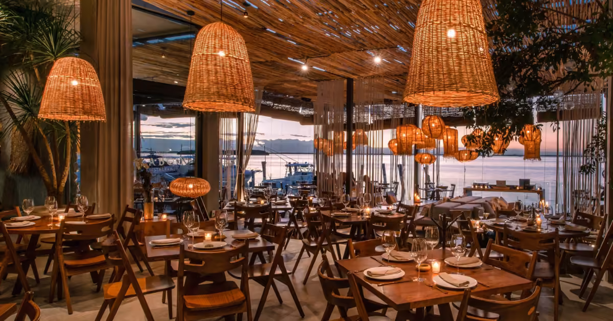 Cozy restaurant with wooden tables, wicker lights, and large windows offering sunset views of a tranquil waterfront.