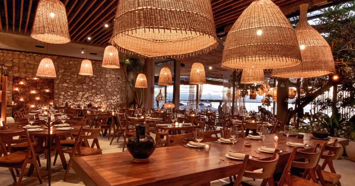Warmly lit restaurant with wooden tables, wicker lamps, stone walls, and large windows framing a scenic sunset; tables set for dining.