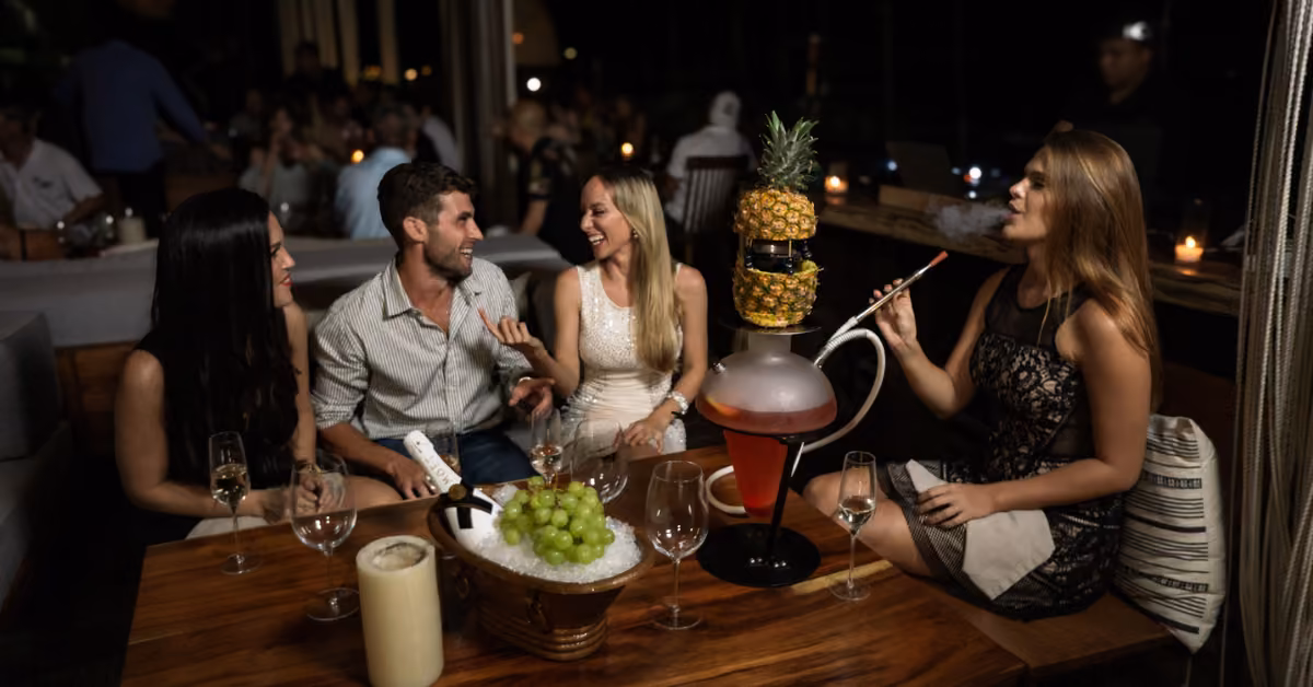 Four friends share drinks and a hookah around a wooden table, laughing together in the cozy, dimly lit lounge.