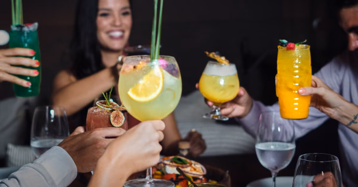 Friends toast with colorful cocktails at a dimly lit table, smiling and celebrating together at Taboo Restaurant.