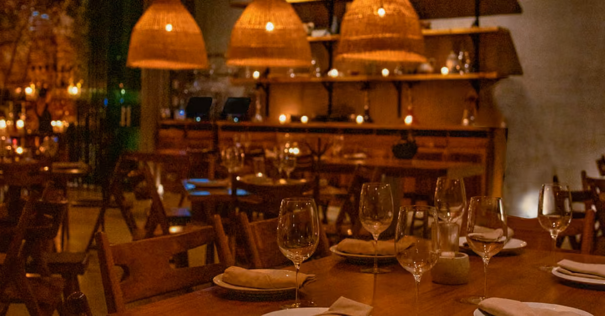 A warmly lit restaurant with wooden tables set for dining and wicker pendant lamps, creating a cozy, inviting atmosphere.