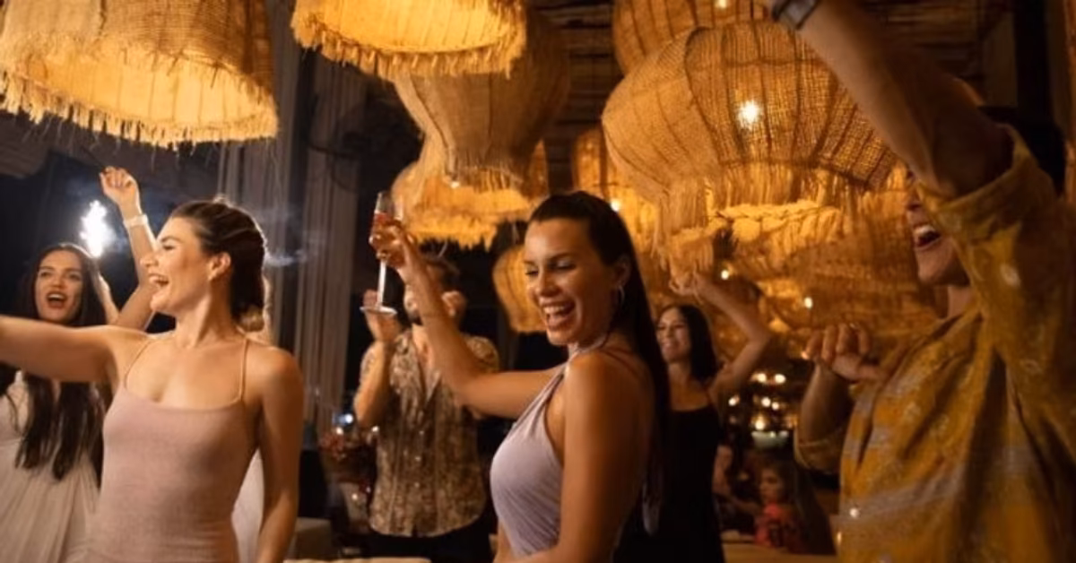 Smiling people toast drinks at a lively indoor party under large lights and cozy, festive decor in a busy restaurant.
