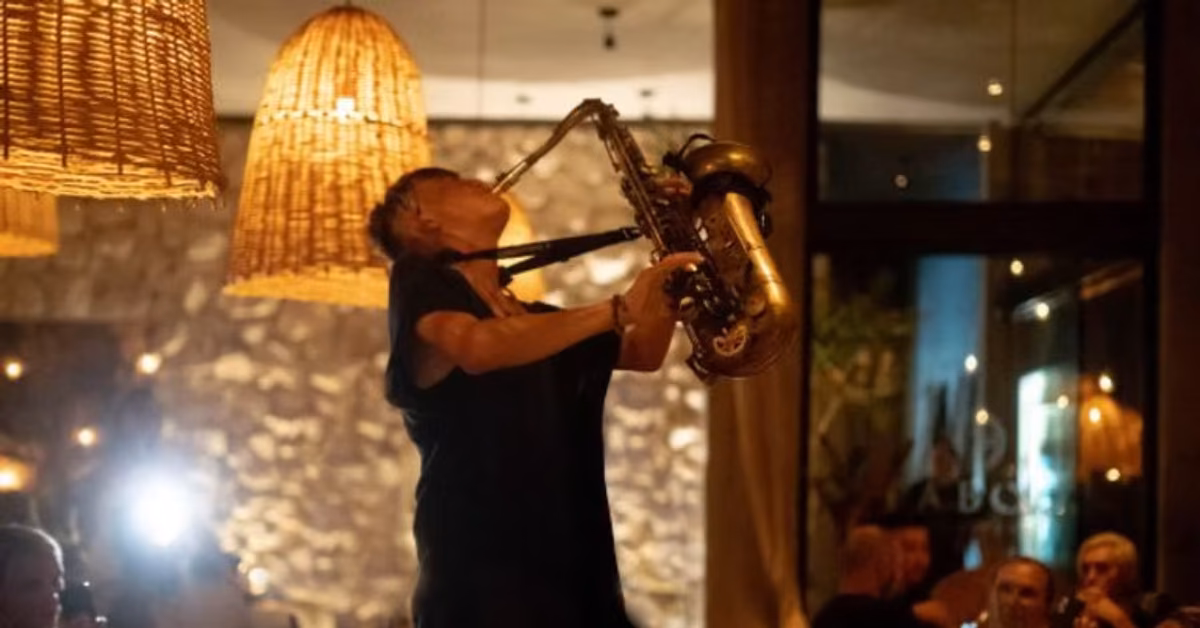 A musician plays saxophone in a warmly lit Taboo Restaurant, surrounded by wicker lamps and an attentive audience.
