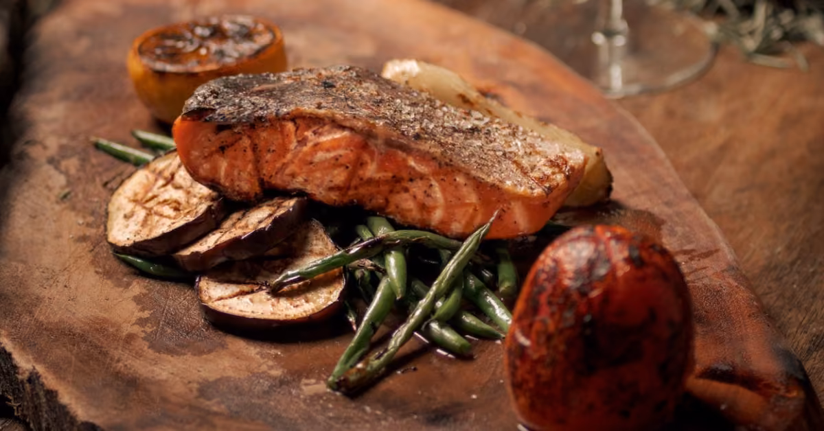 Grilled salmon with roasted green beans, eggplant, and charred tomato, topped with a slice of grilled lemon on a wooden board.