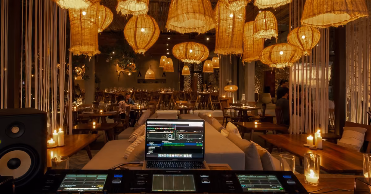Cozy, dim Taboo Restaurant with wicker lamps, wooden tables, and candles. DJ booth with laptop and audio gear in front.