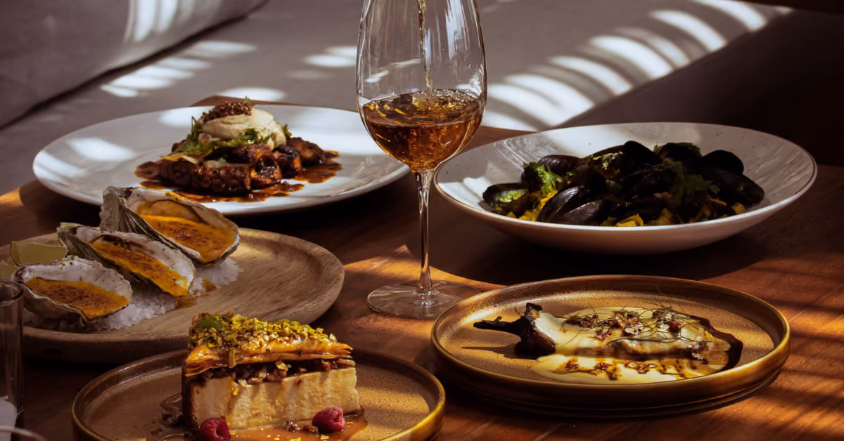 A sunlit table at Taboo Restaurant with oysters, mussels, dessert, and a glass of white wine.