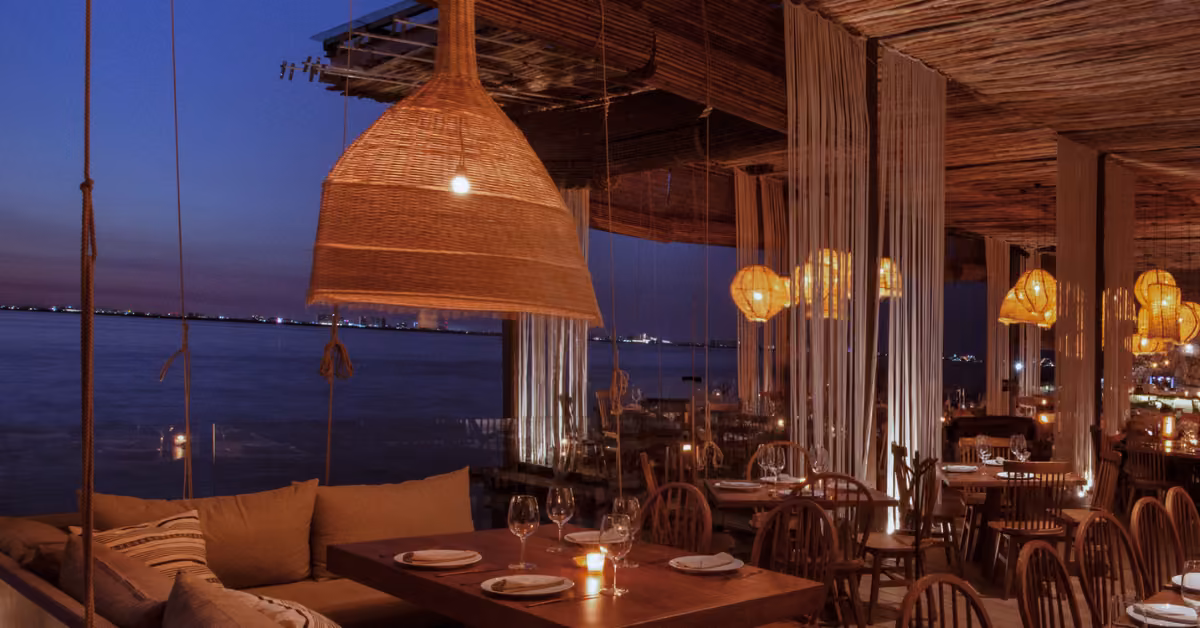 A cozy seaside restaurant at dusk with wooden tables, cushioned seats, candlelight, pendant lights, and ocean views.