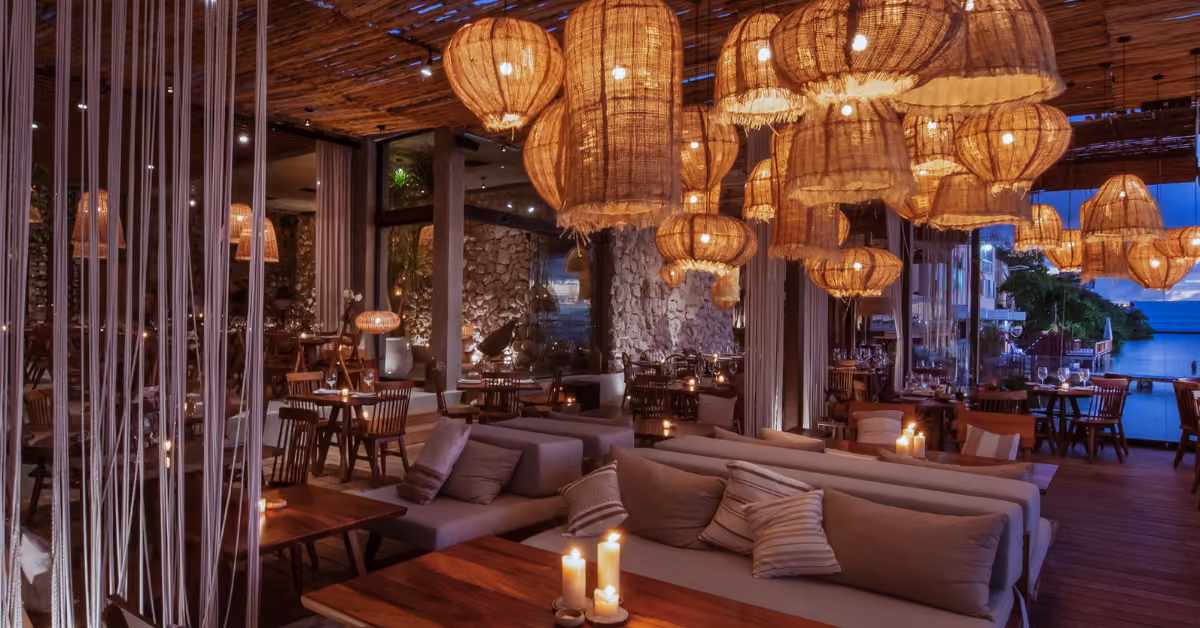 Cozy restaurant with wooden tables, cushioned seats, soft candlelight, and large windows overlooking the sea at dusk.
