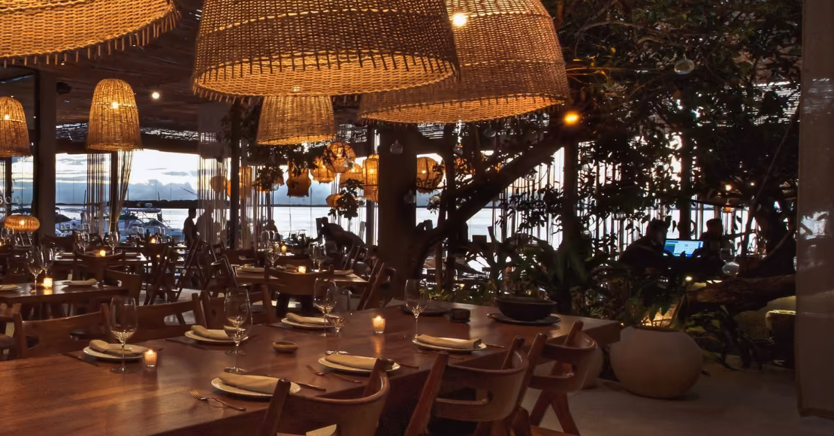 A cozy, softly lit restaurant with wooden tables, wicker lights, candles, plants, and sunset views over the water.