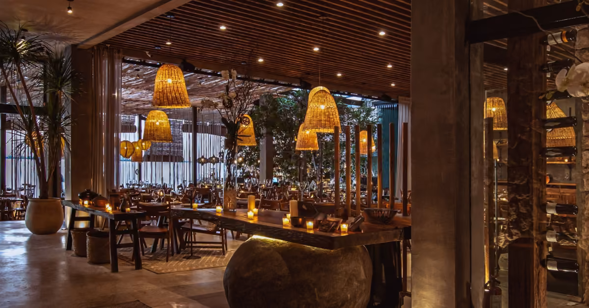 A warmly lit restaurant with wooden tables, wicker lights, potted plants, and candles creates a cozy, inviting atmosphere.
