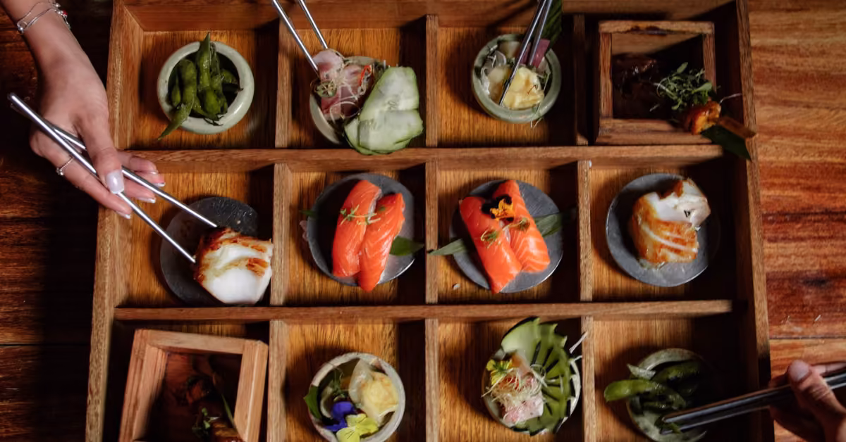 At Tora, two people use chopsticks to pick from a wooden tray of sushi, vegetables, and small dishes on a table.