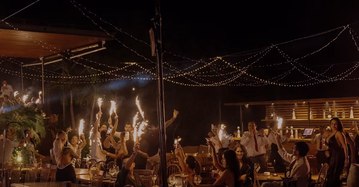 People celebrate outdoors at Tora Restaurant at night, waving sparklers under glowing string lights in a festive scene.