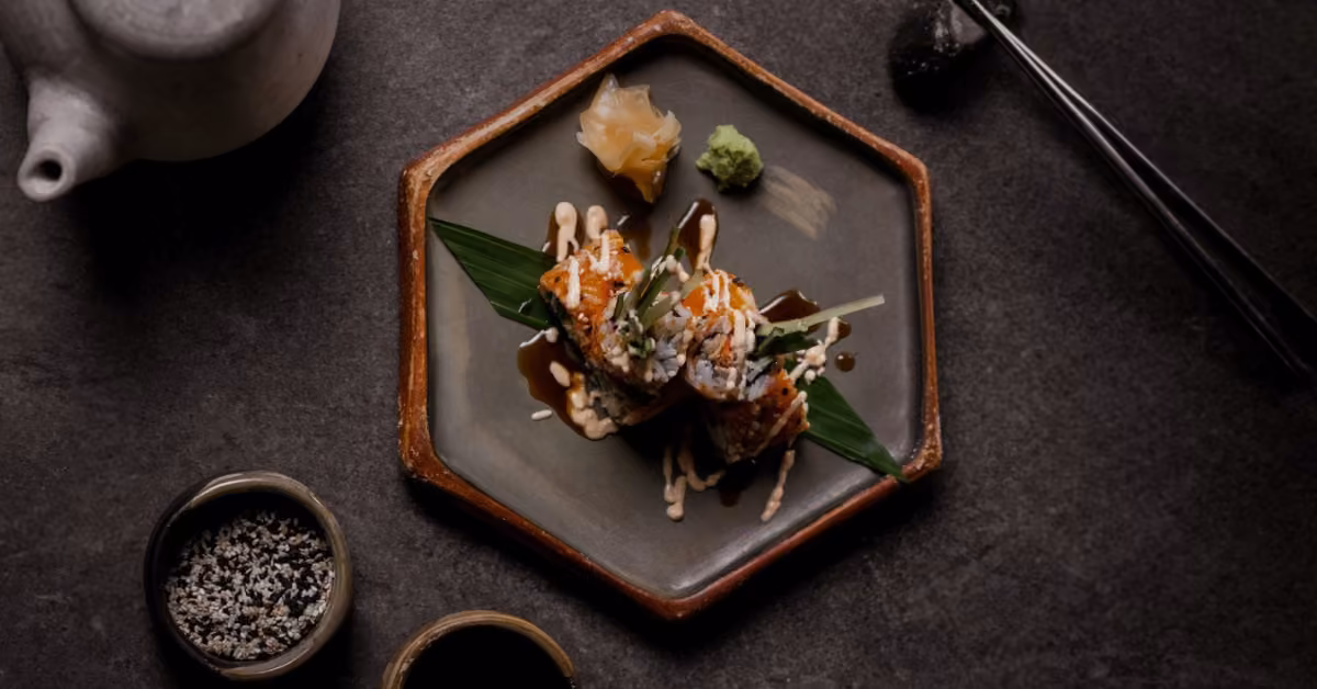 Two sushi pieces with sauce and garnishes on a hexagonal plate, served with wasabi, ginger, chopsticks, and dipping bowls.