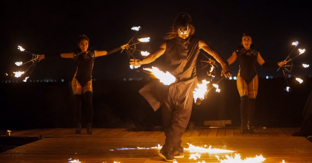 Three performers in black twirl flaming props on a wooden stage at night outside Tora Restaurant, fire illuminating the scene.