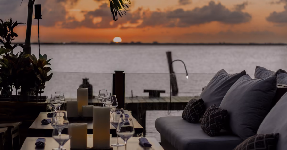 Tora Restaurant’s patio with cushioned seats and candlelit tables, overlooking tranquil water at sunset under an orange sky.