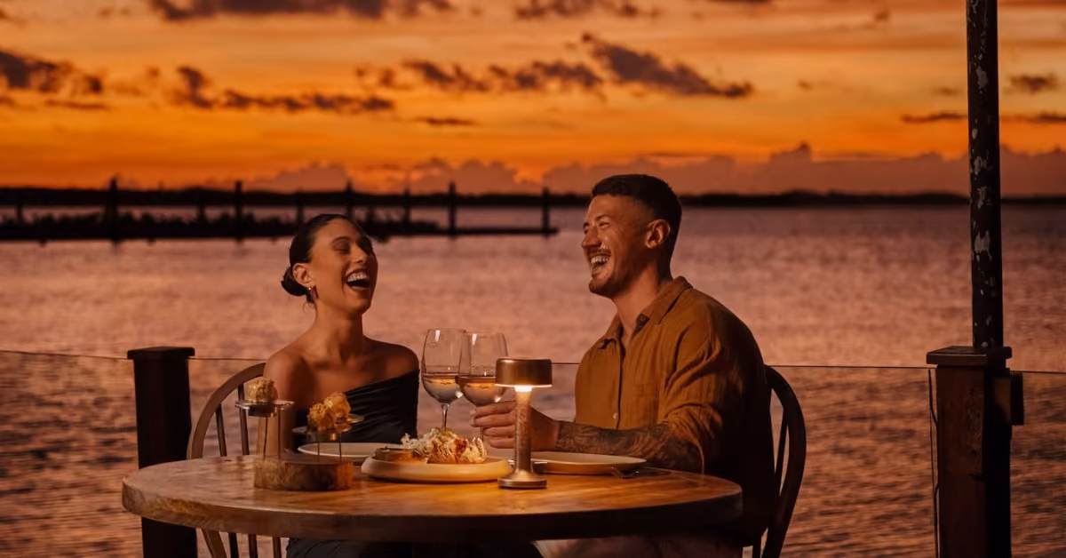 A couple laughs over dinner and wine at a waterfront table during sunset, with boats and a dock in the glowing sky behind them.