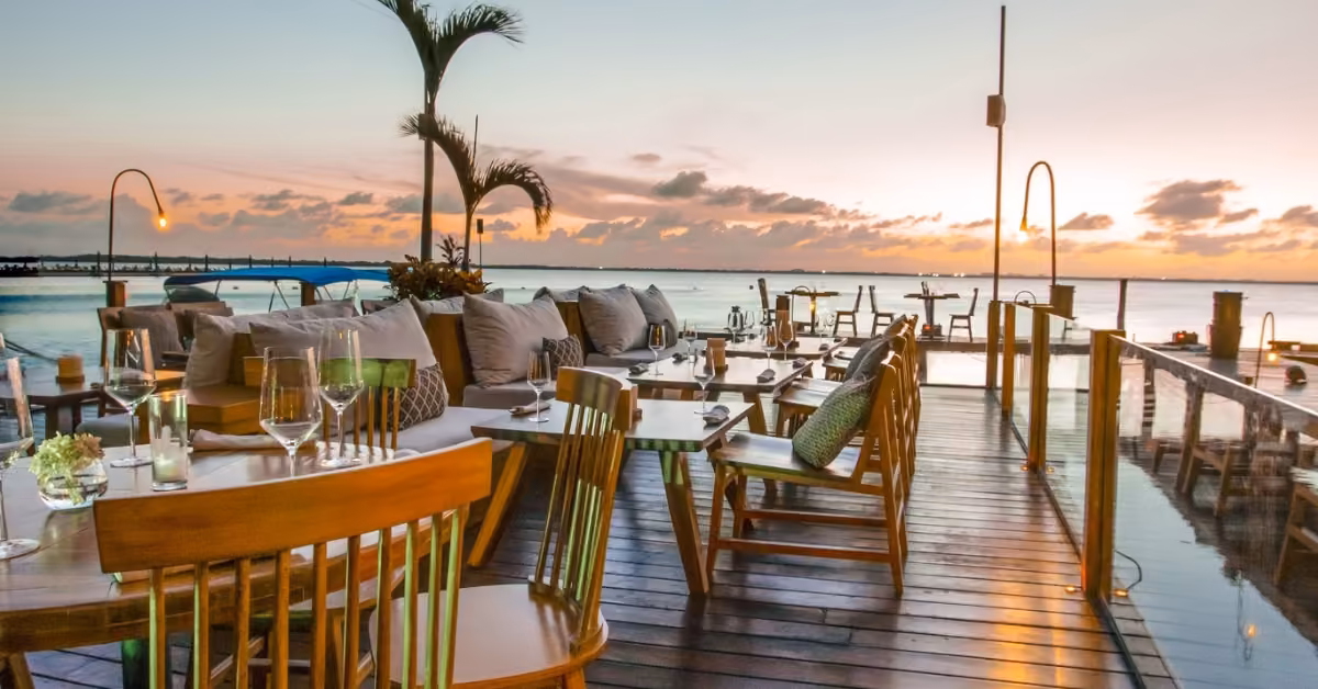 Outdoor wooden tables overlook calm water at sunset, with palm trees and soft lighting creating a relaxed, elegant vibe.
