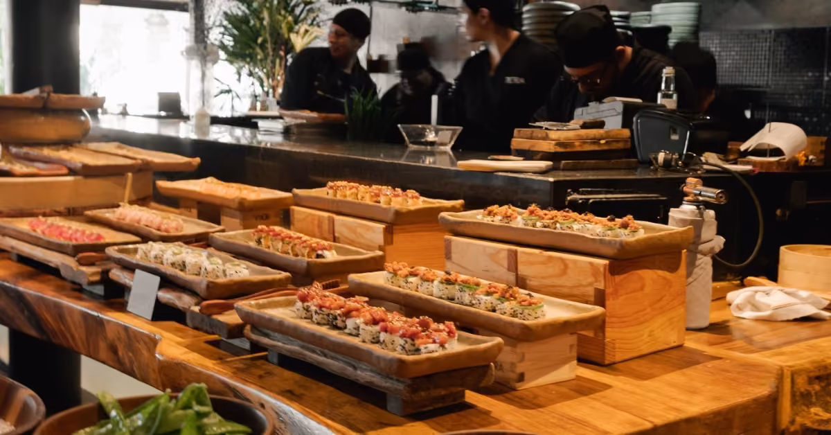 Sushi rolls on wooden trays at Tora Restaurant’s counter, with chefs busy preparing dishes in the background.