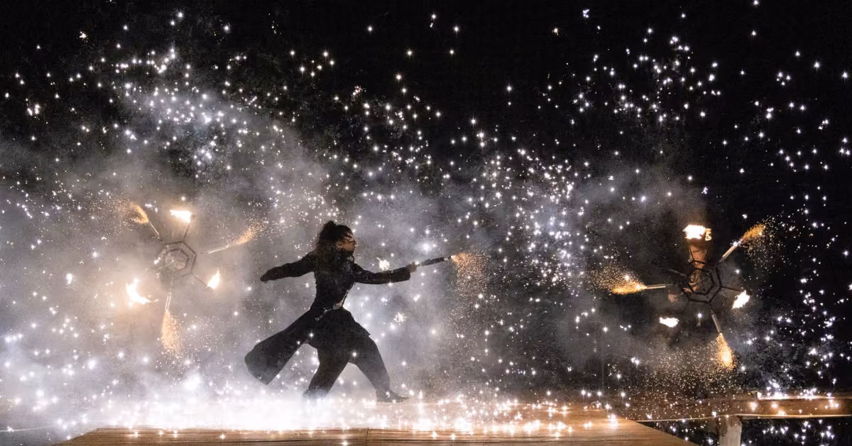 A dancer in dark clothing performs at night outside Tora Restaurant, surrounded by spinning fire and sparkling lights.