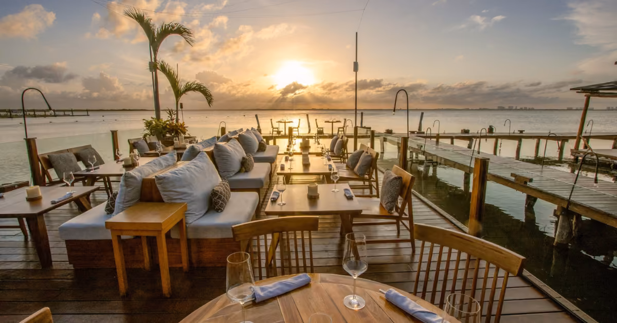 Outdoor restaurant with cushioned seats and wooden tables overlooking the ocean at sunset, creating a warm, relaxing vibe.