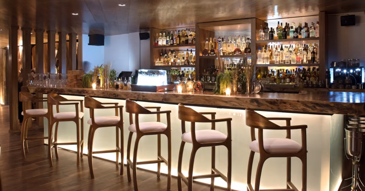 Warmly lit bar at Tora Restaurant with wooden chairs at a glowing counter, shelves of bottles, and elegant, inviting decor.