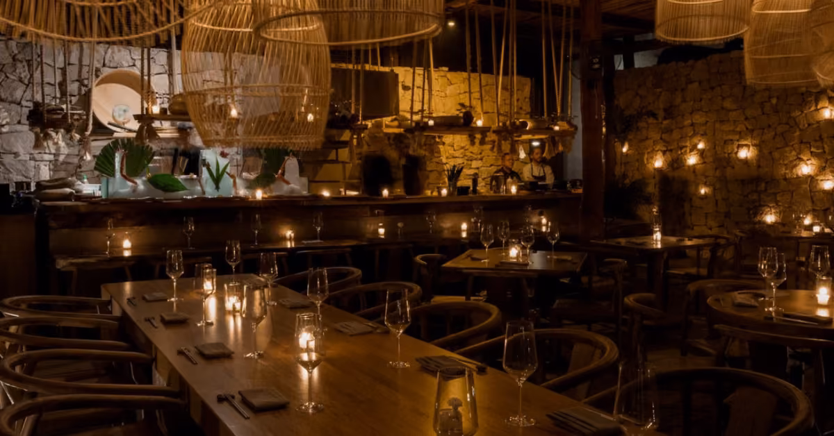 A dimly lit restaurant with wooden tables, wicker lights, candles, stone walls, and an open kitchen for a cozy, rustic feel.