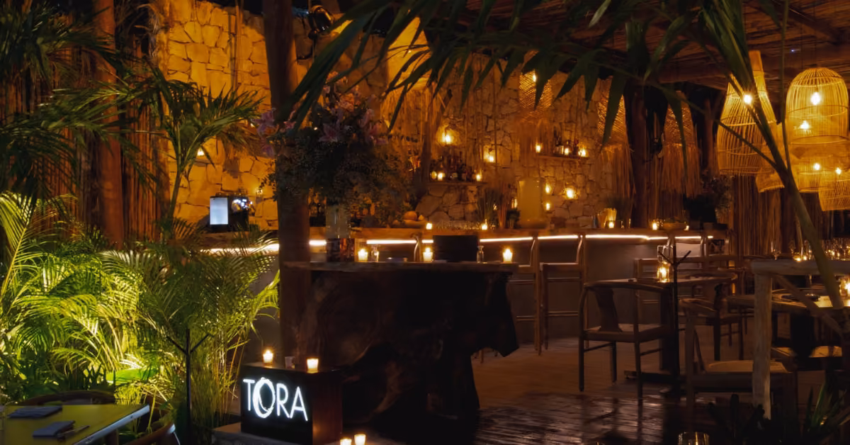 A cozy restaurant with stone walls, rattan lamps, candles, green plants, and a glowing TORA sign in a warm, inviting glow.
