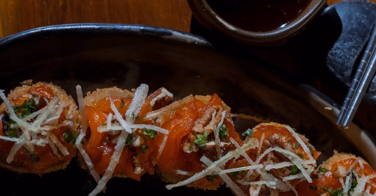 Sushi rolls with sliced salmon, crispy strips, and herbs on a dark plate, served with sauce and chopsticks at Tora Restaurant.
