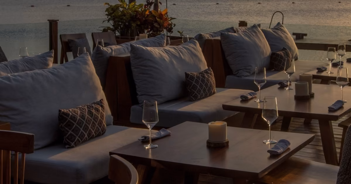 Tora Restaurant’s patio has cushioned sofas, pillows, and wooden tables set for dinner beside calm water at sunset.