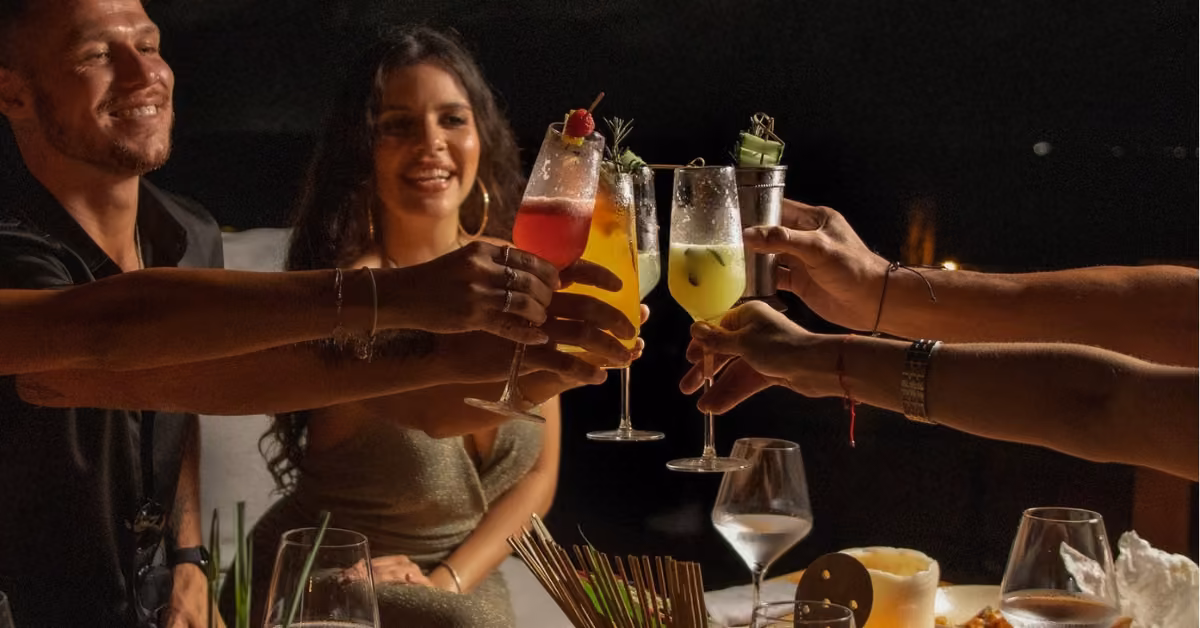 Four friends toast with cocktails, smiling around an outdoor dinner table at night. The vibe is festive and joyful.