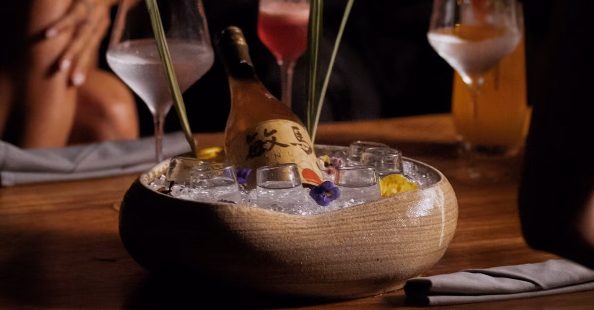 A sake bottle on ice in a ceramic bowl, with shot glasses and flowers on a wooden table at Tora Restaurant.