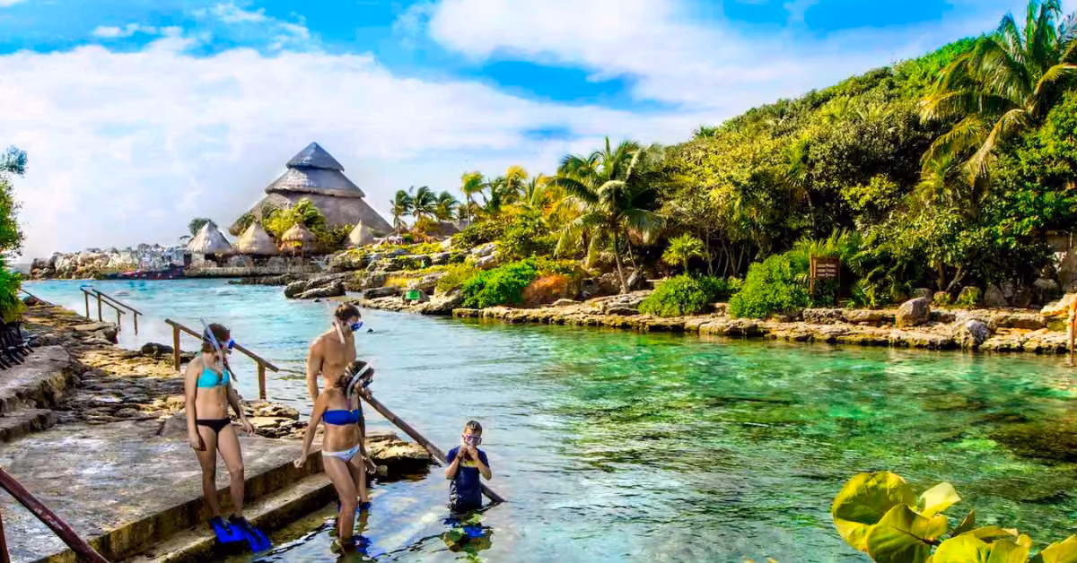 Four people in swimsuits and snorkel gear get ready to enter a turquoise lagoon at Xcaret, surrounded by lush greenery.