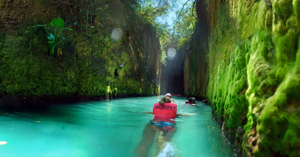 A person in a red life jacket swims through a turquoise waterway at Xcaret, framed by mossy rocks and lush greenery.