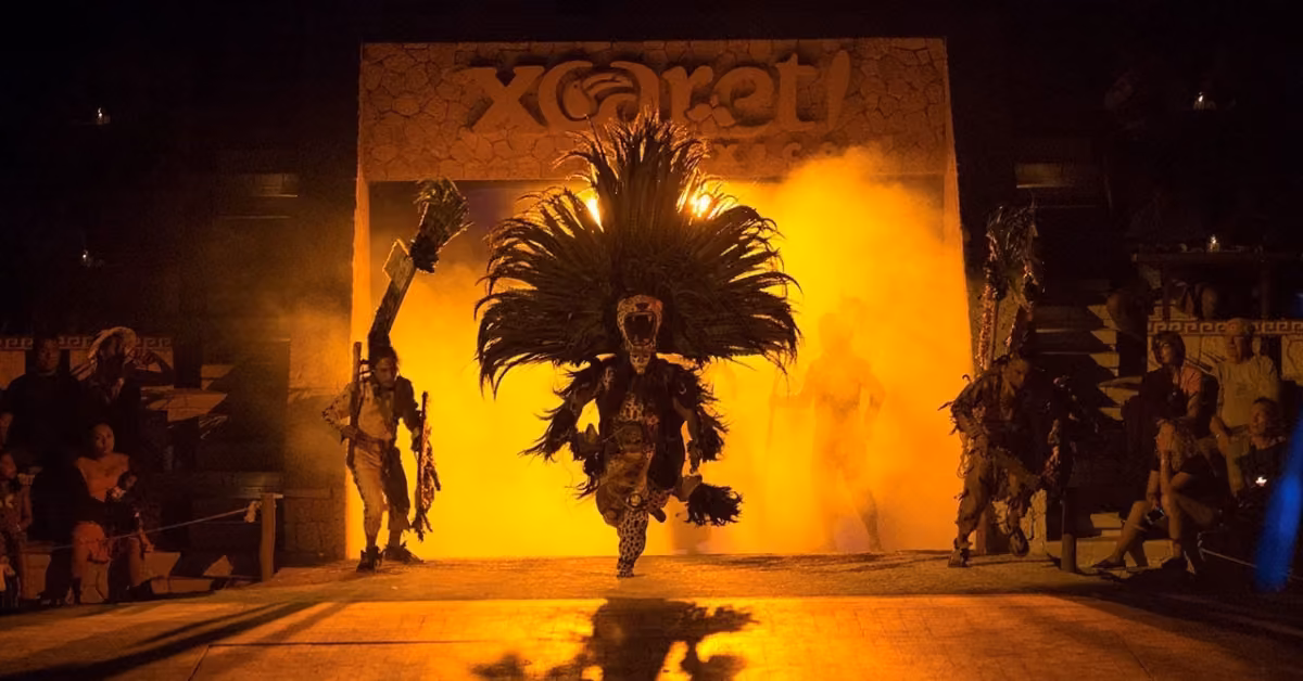 Costumed performers, one in a large feathered headdress, emerge from smoky lights onstage during Xcaret’s vibrant night show.