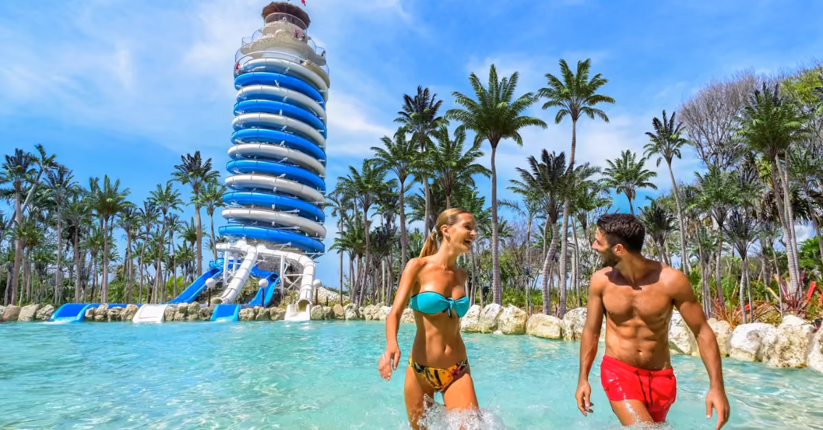 A smiling couple in swimwear walks through shallow water at Xel-Ha, with palm trees and a spiral water slide tower behind them.