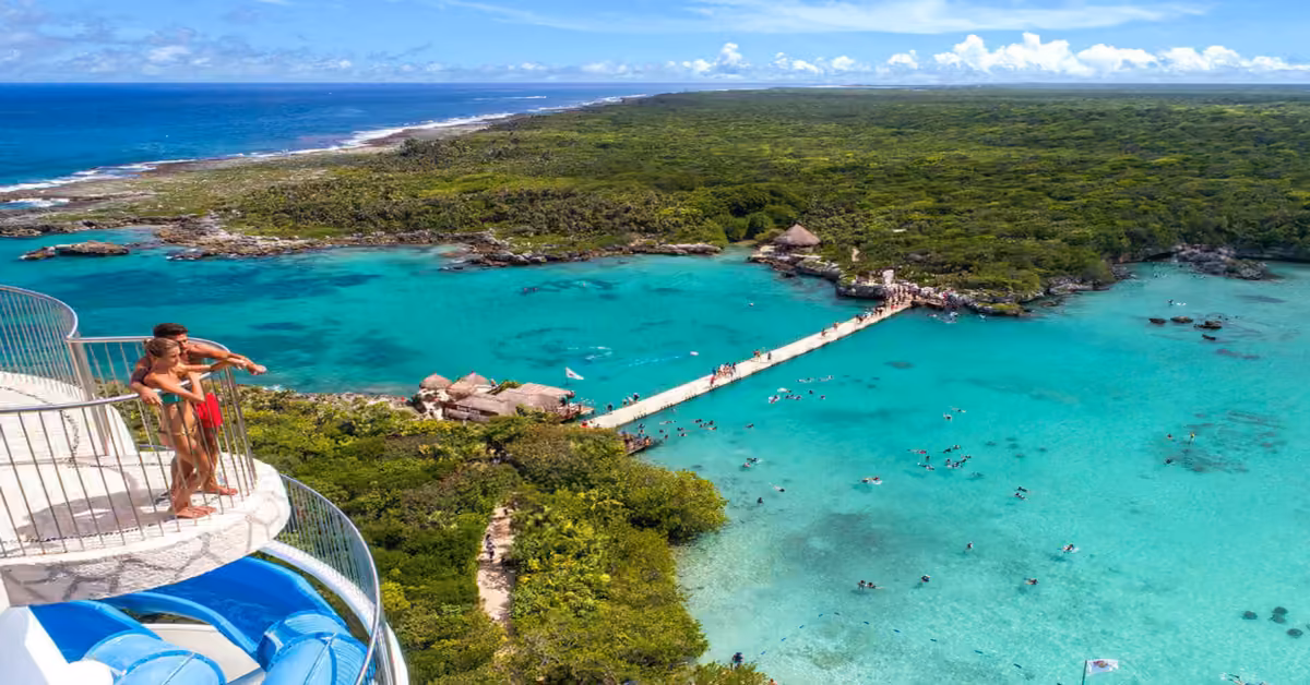 A couple stands on a water slide platform at Xel-Ha, overlooking a turquoise lagoon, lush greenery, and swimmers below.